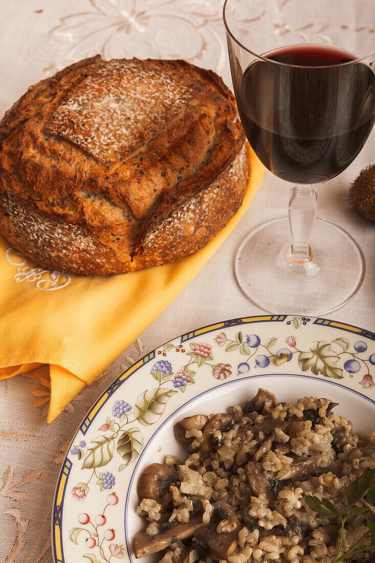 A freshly baked loaf of rustic bread next to a glass of red wine and a plate of savory mushroom risotto on a patterned tablecloth.