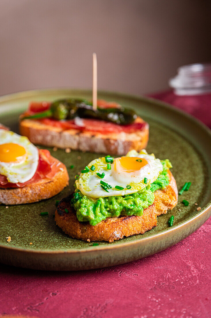 Köstliche hausgemachte spanische Tapas-Toasts mit Spiegeleiern, Avocado und Schinken auf rundem Teller neben knusprigem Brot mit Speck auf dem Tisch serviert