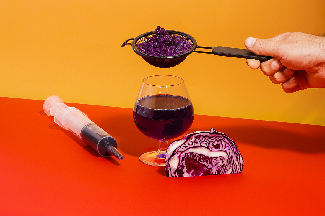 Eine nicht erkennbare Hand hält ein Sieb mit Gemüse, während sie den gesunden, antioxidativen Saft von Rotkohl auf einem Glas auf einem roten Tisch in der Nähe einer Spritze entnimmt