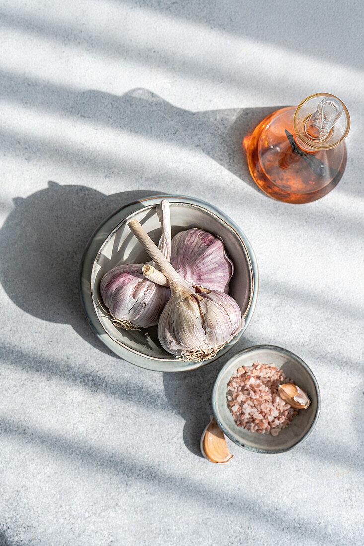 Top view of fresh raw garlic bulbs in a ceramic bowl accompanied by coarse salt and an amber glass bottle all against a textured light gray background