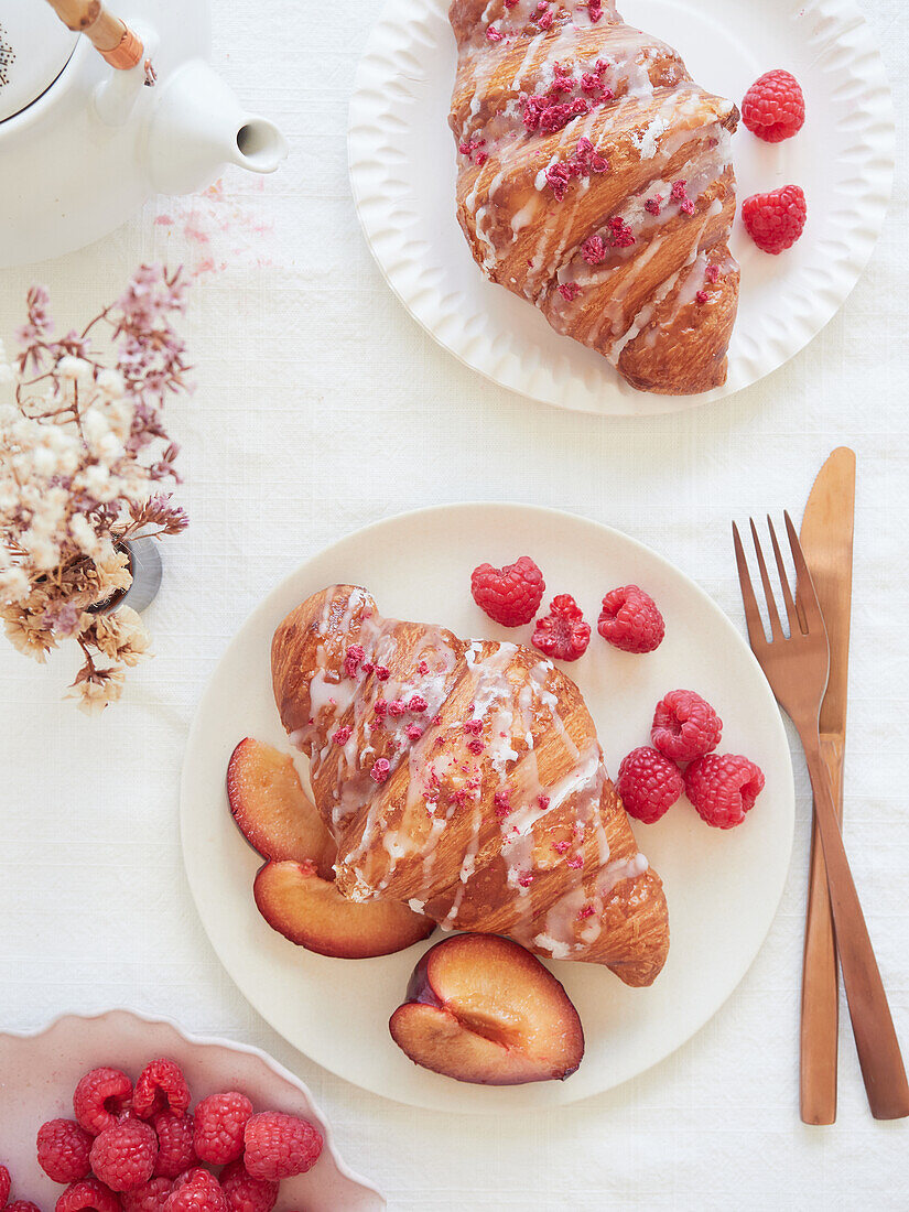 Draufsicht auf ein goldenes Croissant, verziert mit Zuckerguss und bunten Streuseln, umgeben von saftigen Pfirsichscheiben und frischen Himbeeren, serviert auf einem Teller mit Gabel und Messer auf einem Tisch mit Teekanne, Blumen und Himbeeren