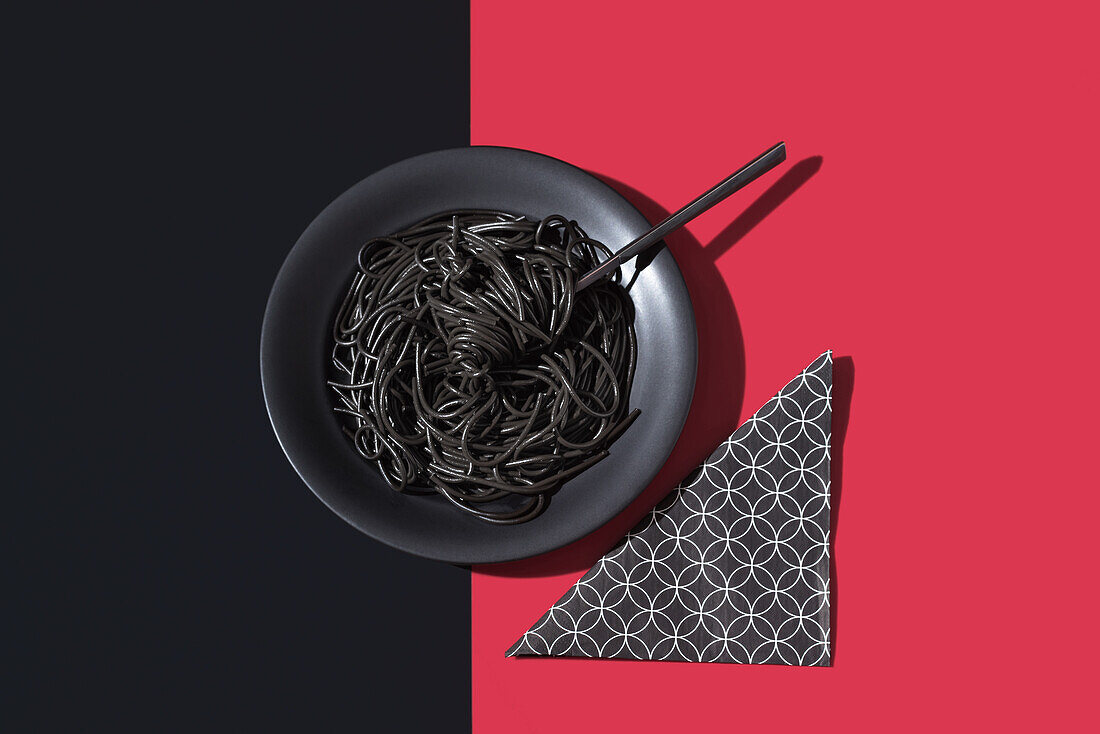 Top view of black spaghetti in a bowl with fork placed on black and red background near napkin