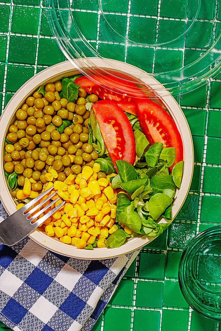 Top view of salad bowl with slices of tomato, spinach leaves, corn kernels and peas placed on napkin on green surface with fork near glass of water
