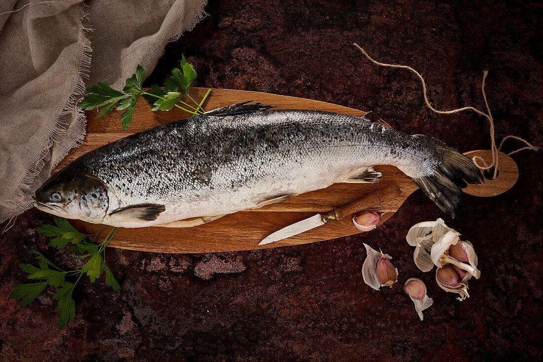 Blick von oben auf ungekochten Fisch mit grünen Blättern und Messer auf einem hölzernen Schneidebrett neben Knoblauch