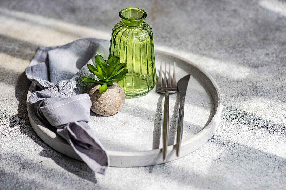 Blick von oben auf einen minimalistisch gedeckten Tisch mit einer kleinen Topfpflanze auf einem Teller mit Besteck und einer leeren Flasche im Sonnenlicht