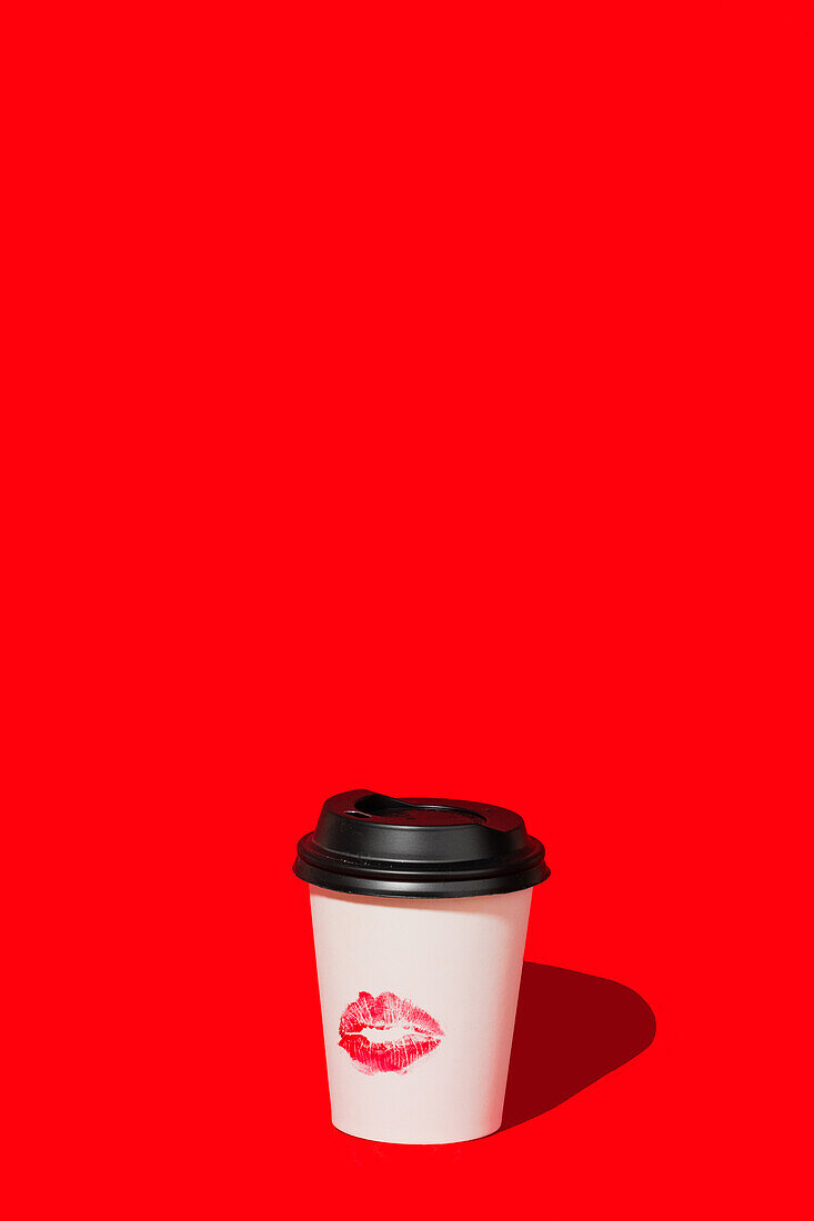 White takeaway coffee cup with a black lid and a lipstick kiss mark isolated against a vivid red background, offering a pop of color.