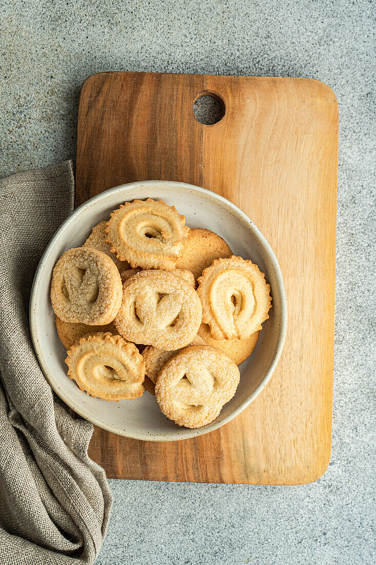 From above fresh homemade baked butter cookies placed in a bowl on a wooden cutting board beside a gray napkin, set against a minimalist concrete background