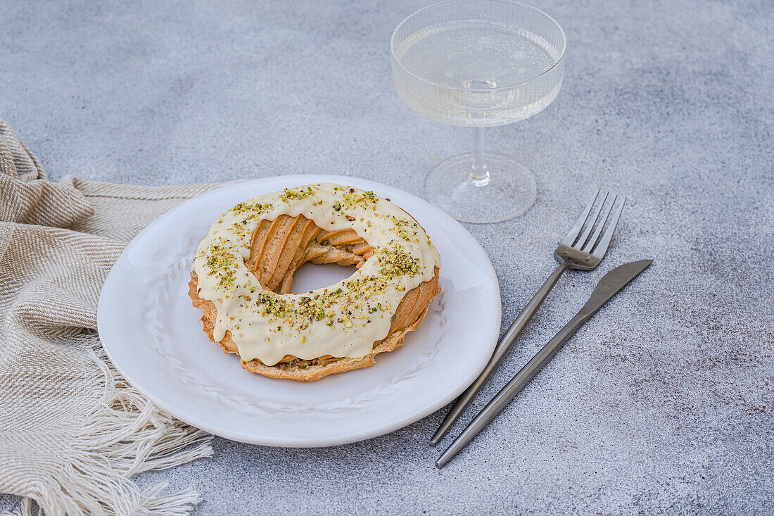 A ring-shaped choux pastry topped with white icing and sprinkled with chopped pistachios, presented on a white plate with a fork and glass, placed on a beige placemat over a textured grey surface