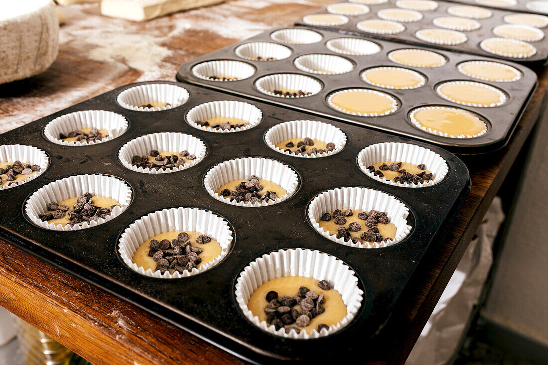 Nahaufnahme eines metallischen Muffinblechs mit Cupcake-Teig und Schokoladenstückchen in Papierformen, die zum Backen auf einem Tisch in einer Backstube angeordnet sind