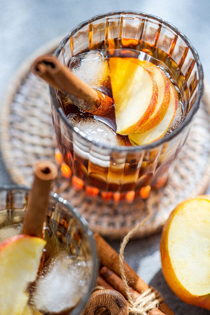 Top view of fresh apple cider cocktails garnished with cinnamon sticks, star anise, and apple slices in a crystal glass filled with ice cubes on a textured background