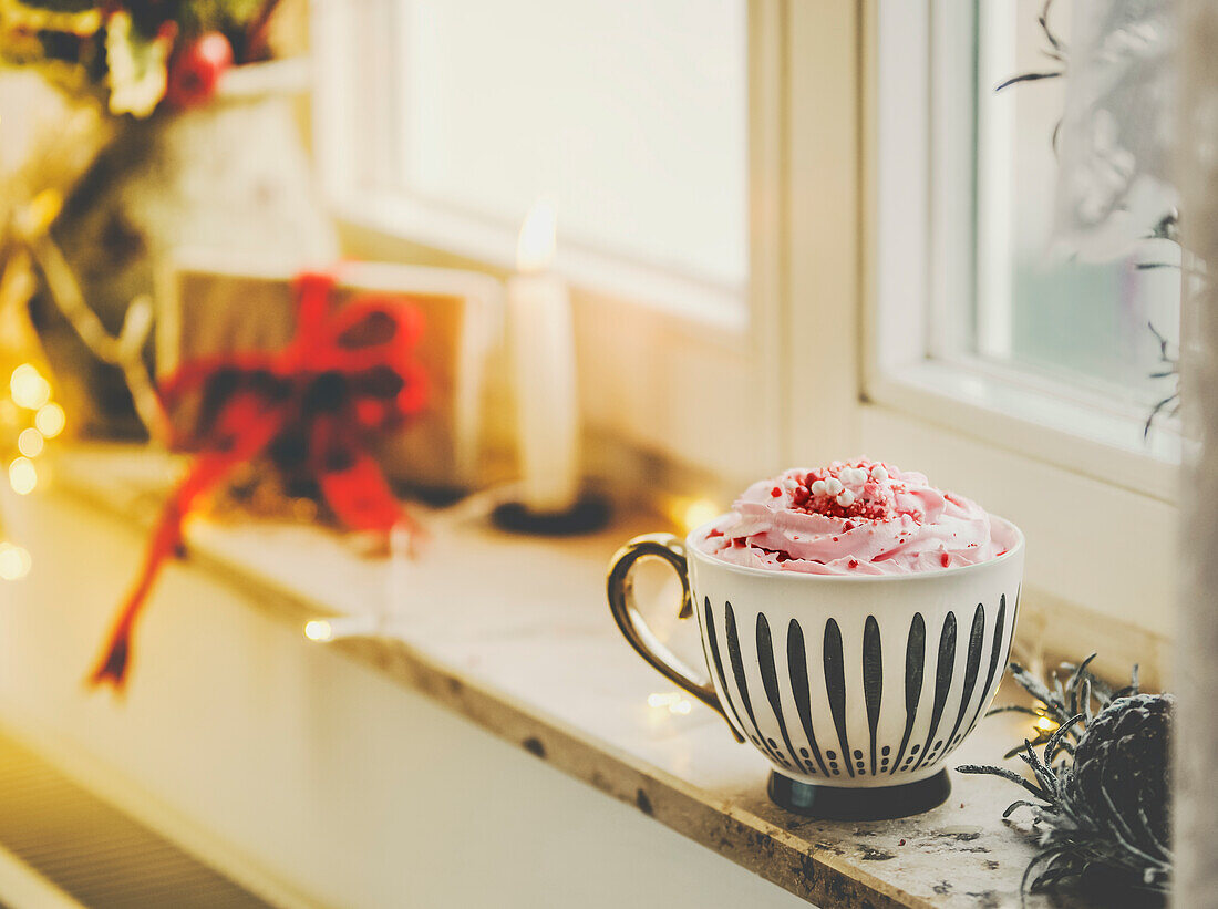 Mug with hot chocolate and whipped cream on window sill with candle, present and warm light. Hygge winter at home with warming drink.
