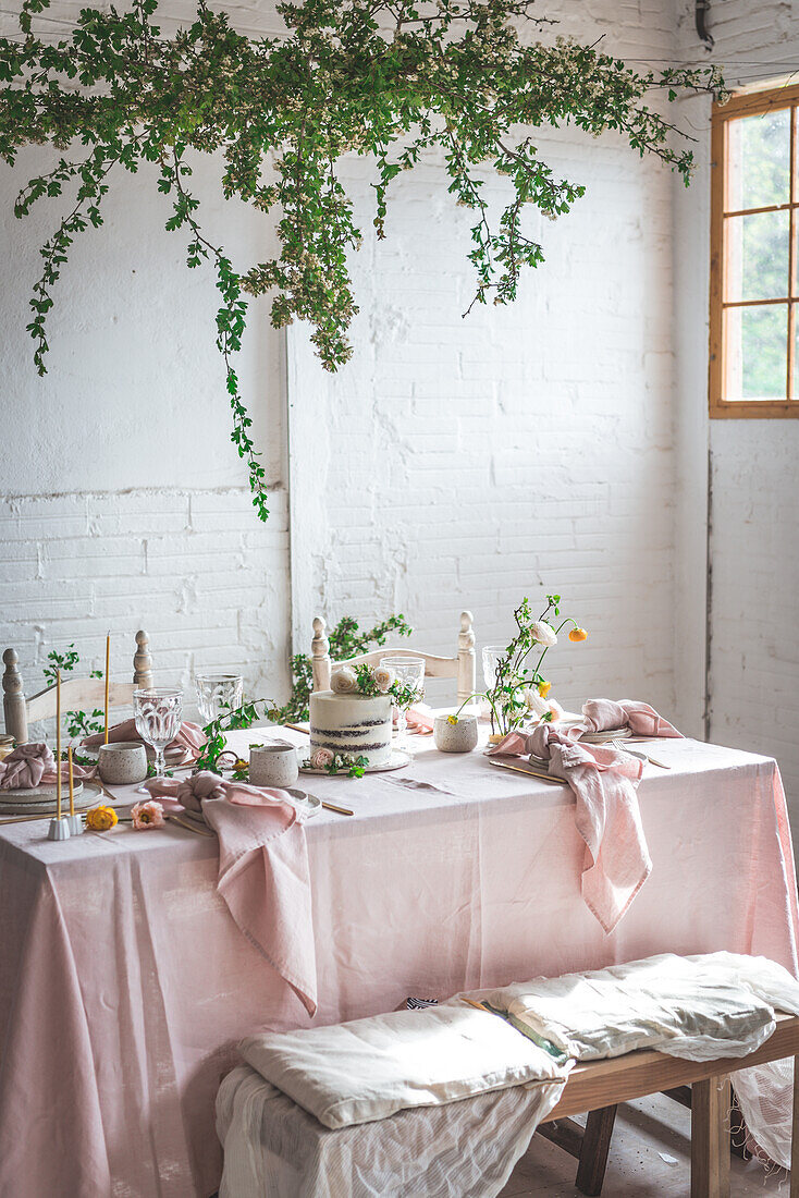 Elegant table served with plates and flowers placed near yummy cake on pink tablecloth against brick wall