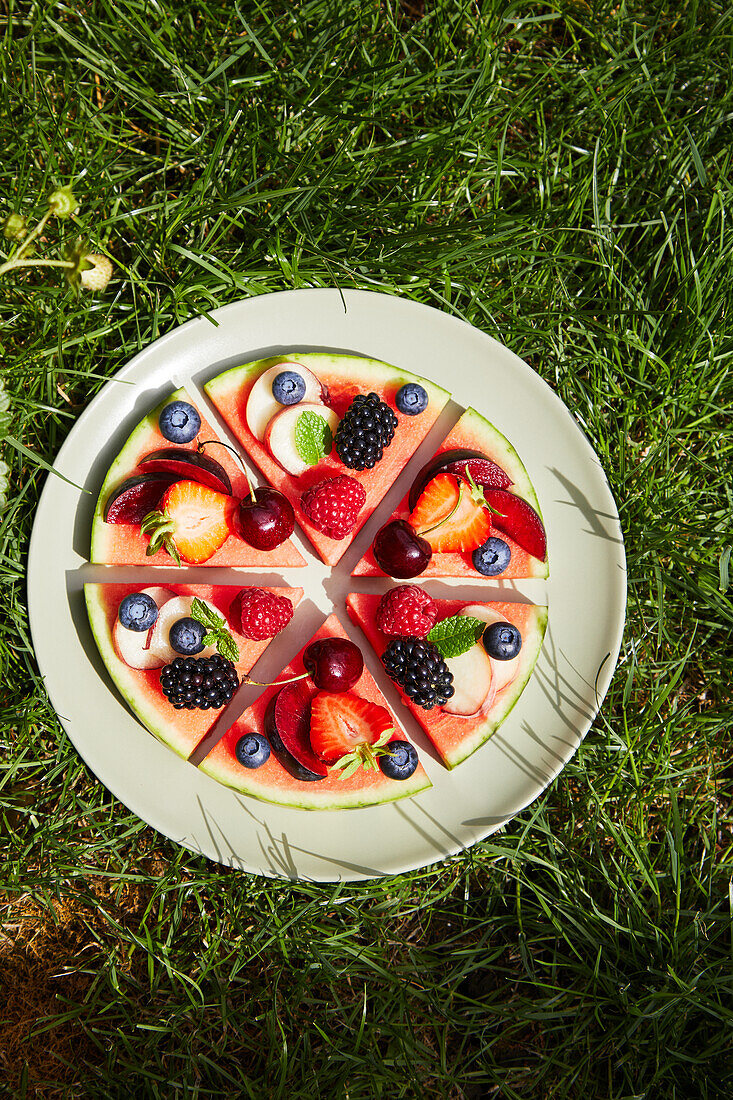 Draufsicht auf Zutaten, bestehend aus Beeren, Äpfeln, Weintrauben, Pflaumen und Pfirsichen, die auf rund geschnittenen Scheiben in einem grauen Keramikteller im Sonnenlicht auf einer grünen Rasenfläche platziert sind, während sie eine Wassermelonenpizza zubereiten