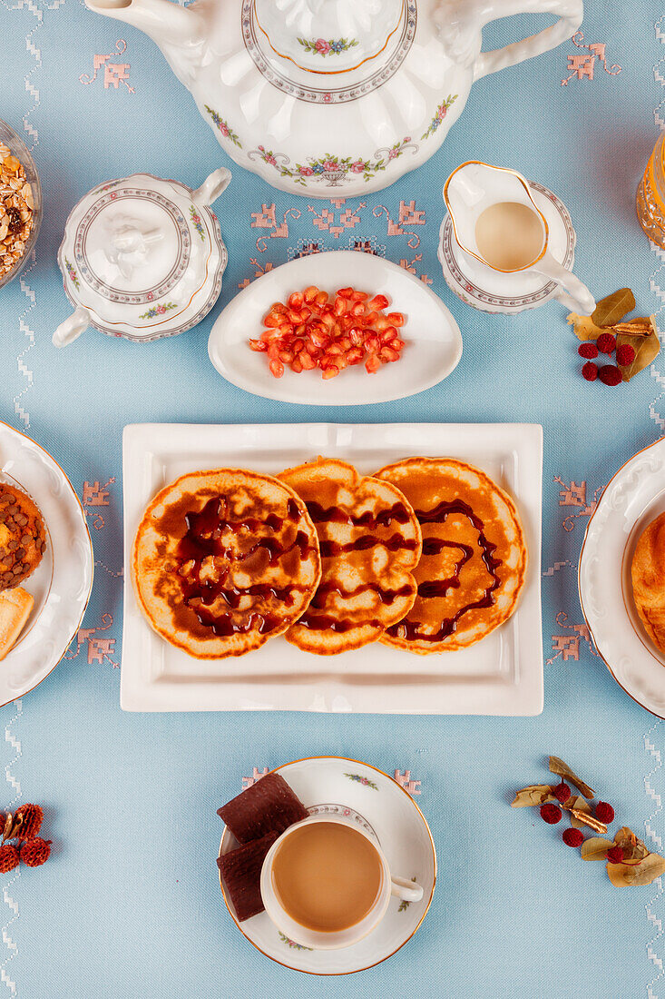 A top view of an afternoon tea arrangement with waffles, sweets, and a porcelain teapot on a light blue background.