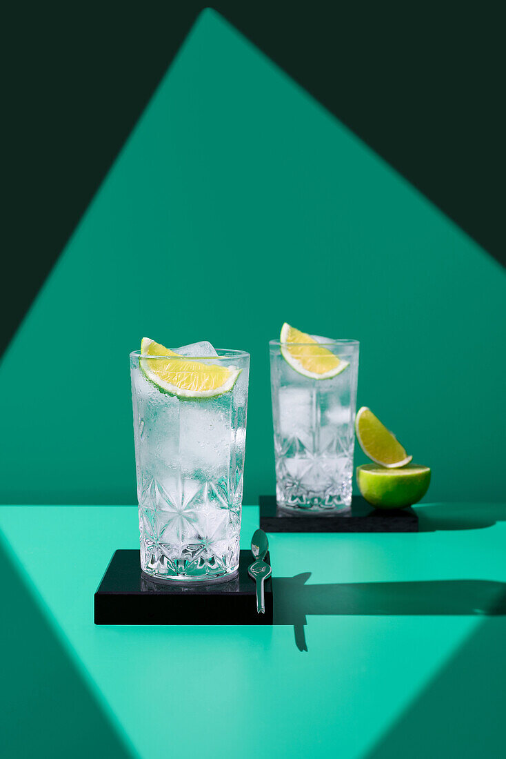 Zwei hohe Gläser, gefüllt mit Gin Tonic und einer Limettenscheibe, präsentiert auf einem minimalistischen grünen Hintergrund mit scharfen Schatten
