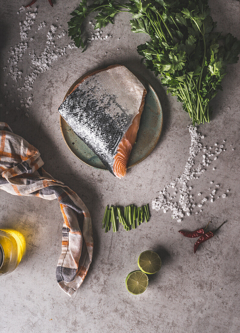 Salmon cooking preparation: whole raw salmon steak with scales at grey concrete kitchen table with herbs, salt, lime, chili, oil and dish cloth. Preparing dinner at home with fresh fish. Top view.