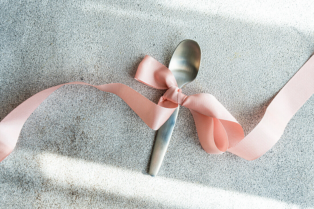 Ein silberner Löffel, der mit einem zarten rosa Band gebunden ist, liegt auf einem strukturierten grauen Tuch und stellt ein einfaches, aber elegantes Tischdetail dar