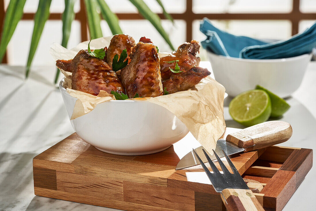 Tasty grilled marinated chicken wings placed in bowl and served with sliced lime on wooden cutting board with knife on table against blurred background in daylight