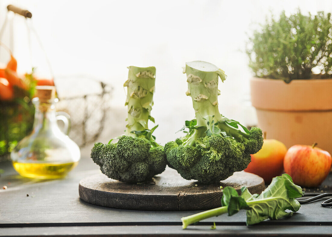Zwei ganze Brokkoliköpfe auf dem Küchentisch mit Zutaten: Olivenöl, getöpfertes Kraut am Fensterhintergrund mit natürlichem Licht. Kochvorbereitung mit gesundem grünem Gemüse. Ansicht von oben