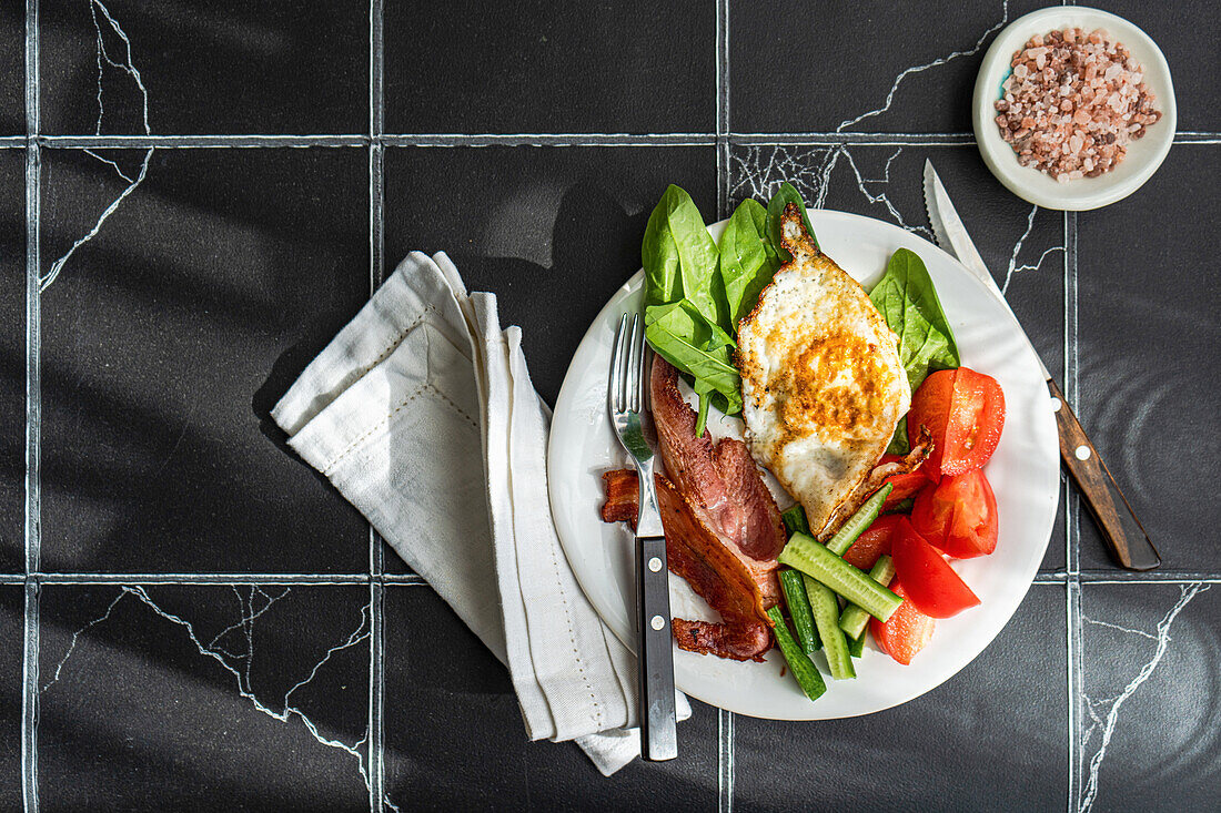 Top view of well balanced breakfast setup with a fried egg, bacon strips, and assorted vegetables like tomato, cucumber, and spinach on a white plate over a dark marble background