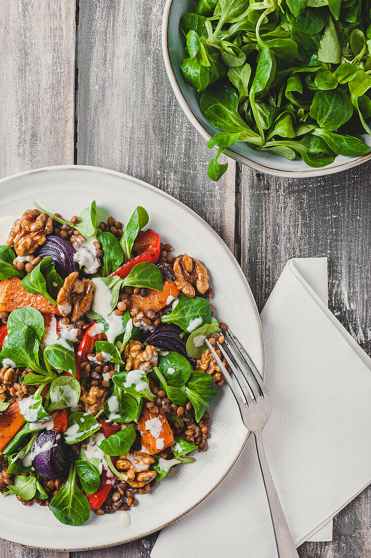 Top view of appetizing fresh salad with green leaves and walnuts seasoned with sauce and served on white plate with fork near bowl with spinach