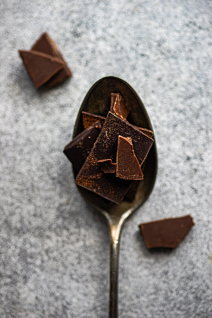 Draufsicht auf Stücke verschiedener Schokoladensorten, die auf einem Erntelöffel und auf einem grauen, unscharfen Tisch liegen