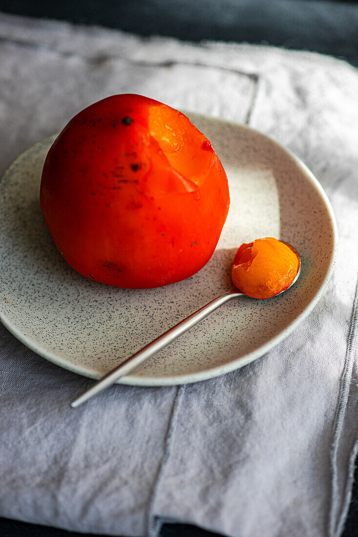 A vibrant, ripe persimmon rests on a ceramic plate beside a spoon, with dramatic shadows accentuating its curves.