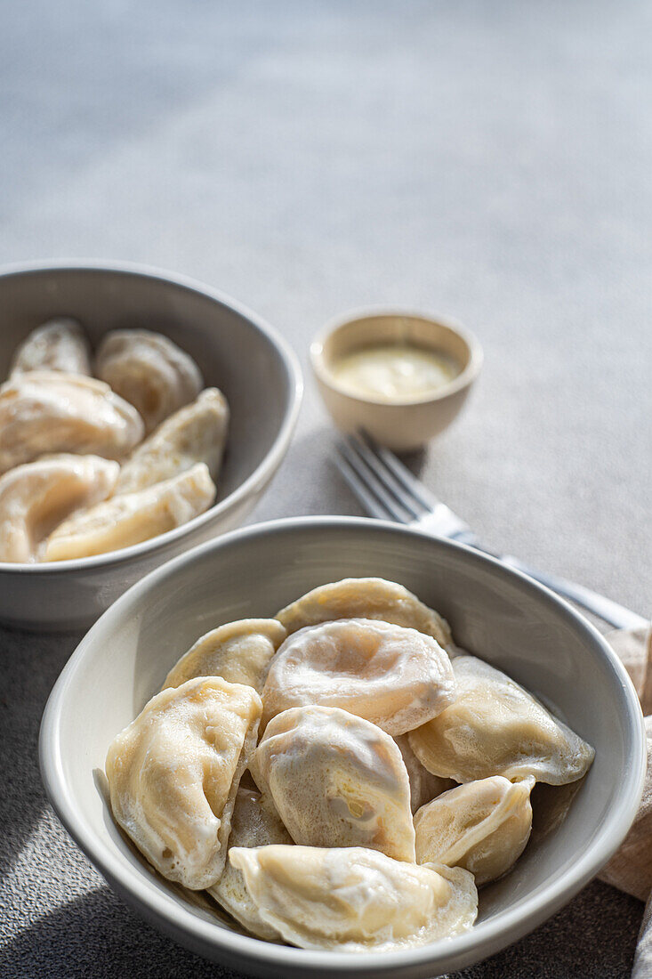 Freshly prepared Ukrainian varenyky dumplings in a white bowl with a side of sour cream and butter, on a textured grey surface with a fork