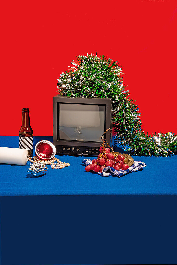 Vintage television set surrounded by an array of objects, including a bottle with a striped label, fresh grapes on a checkered cloth, a white candle and green tinsel, all set against a red backdrop on a blue table