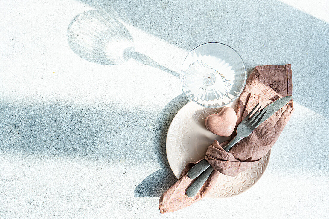 Top view of ceramic plate with cutlery and napkin with heart shaped decor crystal placed on concrete surface at kitchen table for meal during Valentine's day celebration