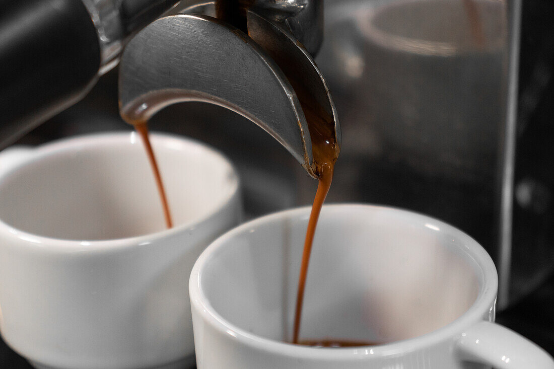 Professional coffee maker pouring hot drink into in two white cups on blurred background