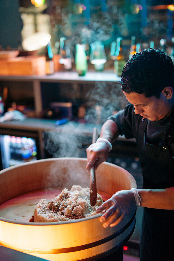 A sushi chef is stirring rice in a wooden tub with a paddle, surrounded by a misty ambiance that suggests a bustling, authentic sushi kitchen.