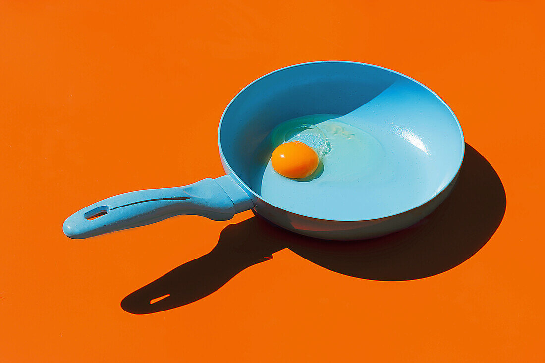 Whole raw egg yolk placed in blue frying pan with handle isolated against orange background
