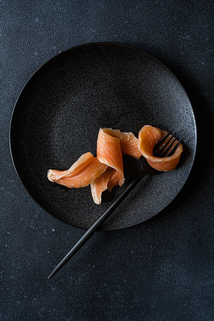 Top view of healthy salmon slice served on black plate near fork against dark surface