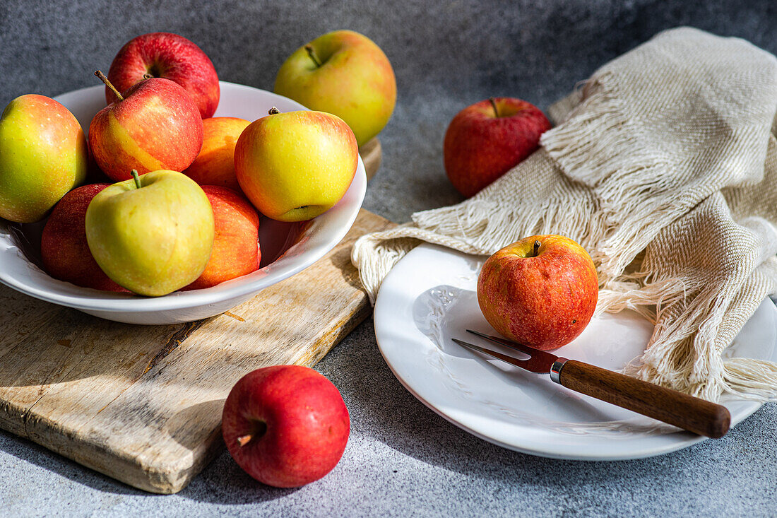 A collection of ripe, colorful apples presented on a rustic wooden board, with a white plate, fork, and draped fabric creating a serene kitchen setting