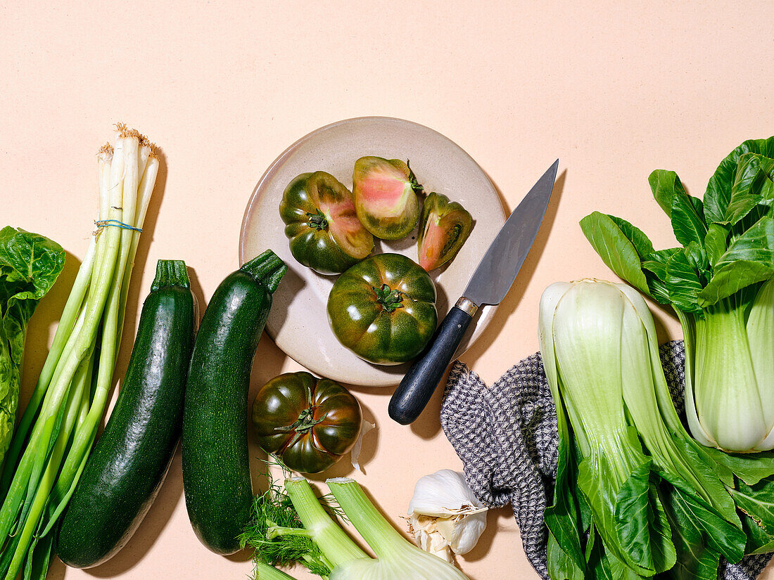 Grüne Tomaten mit anderen Gemüsesorten wie Zucchini und Bok Choy in hellem Sonnenlicht, flachgelegter Teller mit Messer und Kopierbereich auf beigem Hintergrund