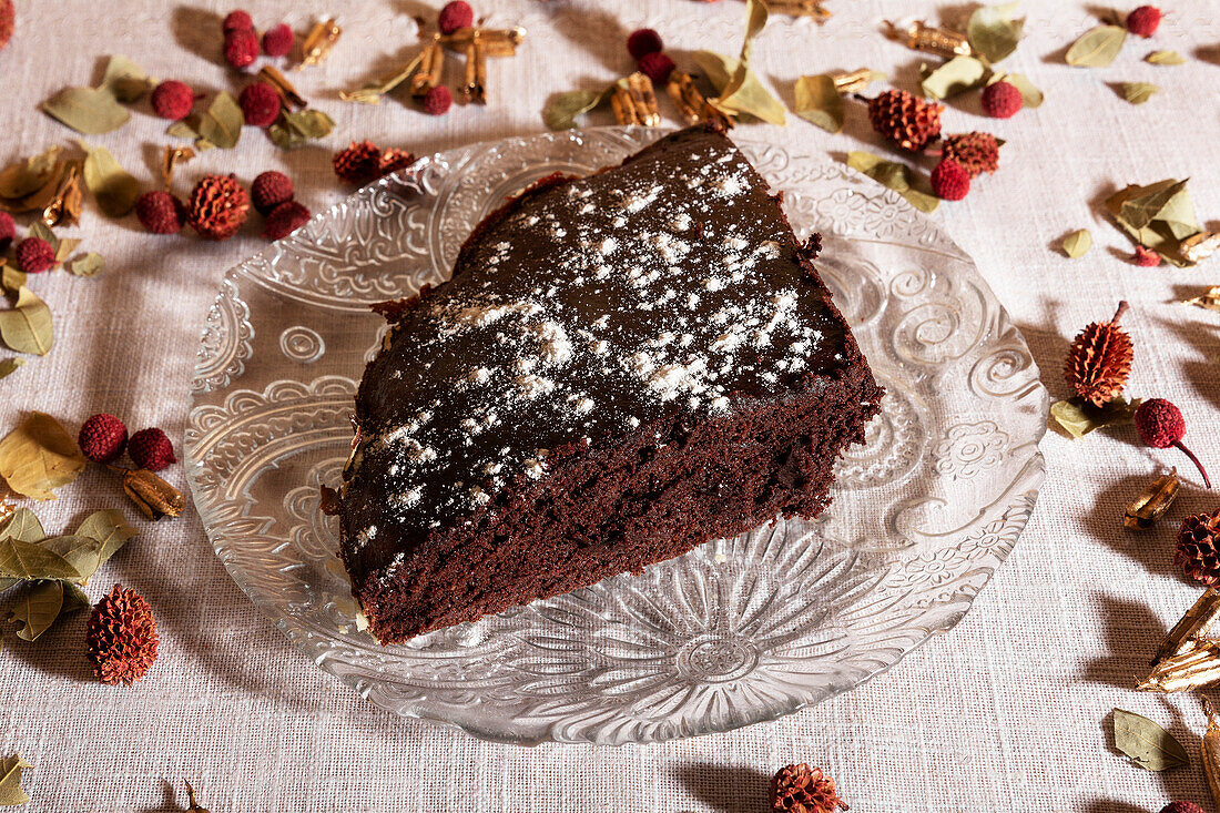 High angle of a slice of rich chocolate cake dusted with powdered sugar on a decorative glass plate, surrounded by dried flowers