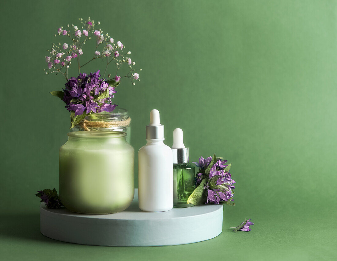 Naturkosmetik mit Pipettenflasche, Kerze und Blumen auf einem Podest vor grünem Hintergrund. Gesundes Hautpflegekonzept mit natürlichen Produkten für die Gesichtsbehandlung. Vorderansicht