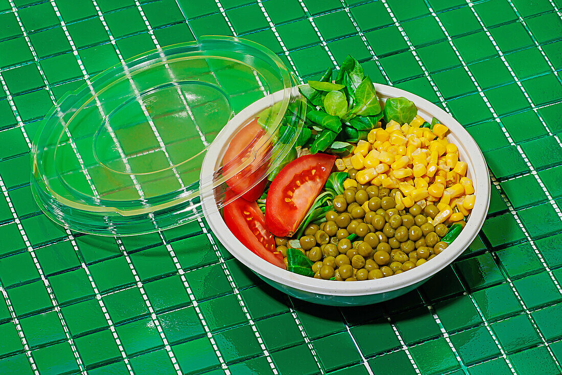 High angle of salad bowl with slices of tomato, spinach leaves, corn kernels and peas placed on green surface
