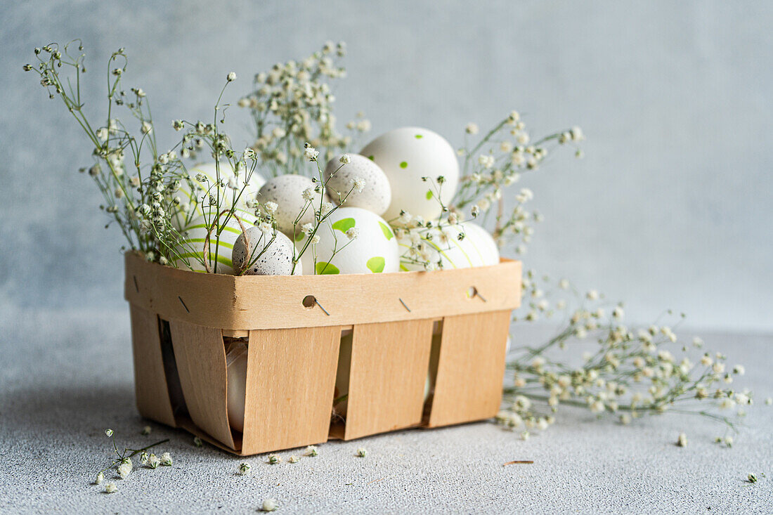 A wooden basket filled with decorated Easter eggs among delicate white spring flowers, on a neutral background