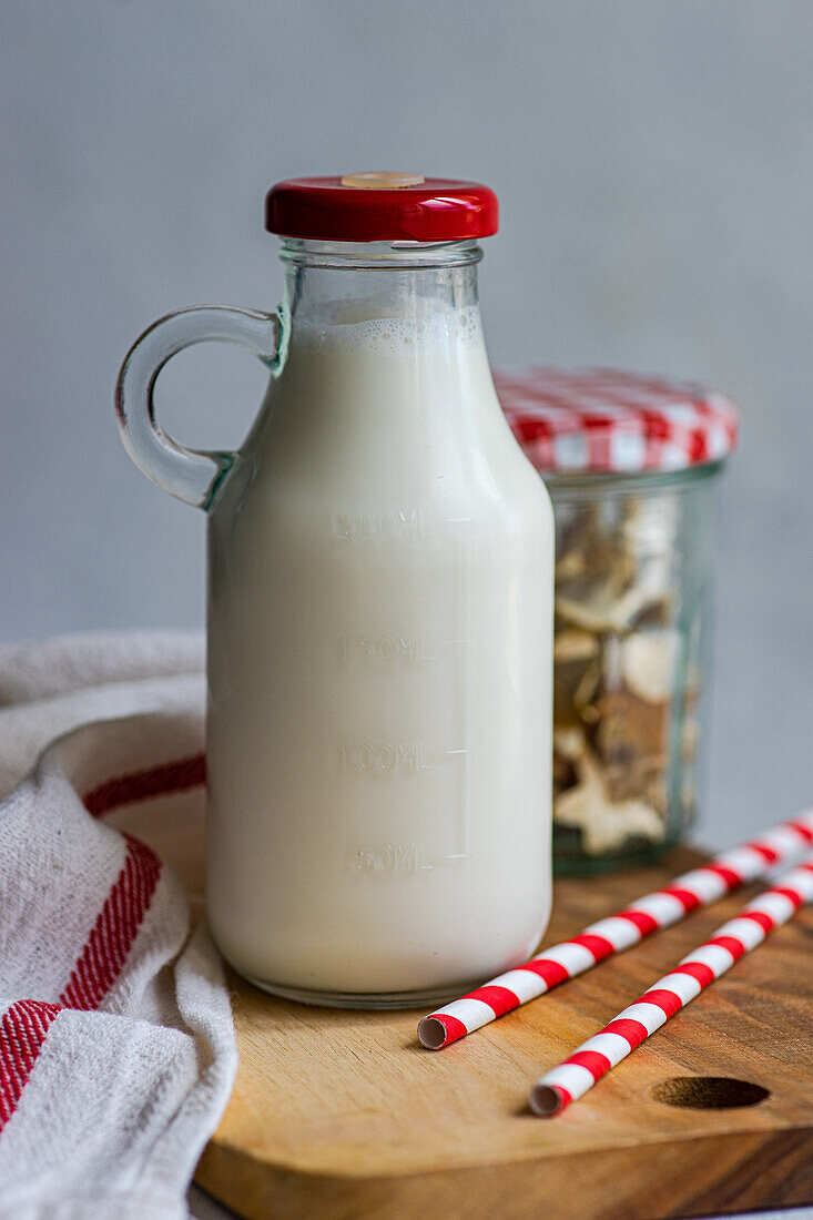 Raw cow milk in vintage closed bottle on cutting board near drinking straws, bottle with cookies and napkin against gray surface