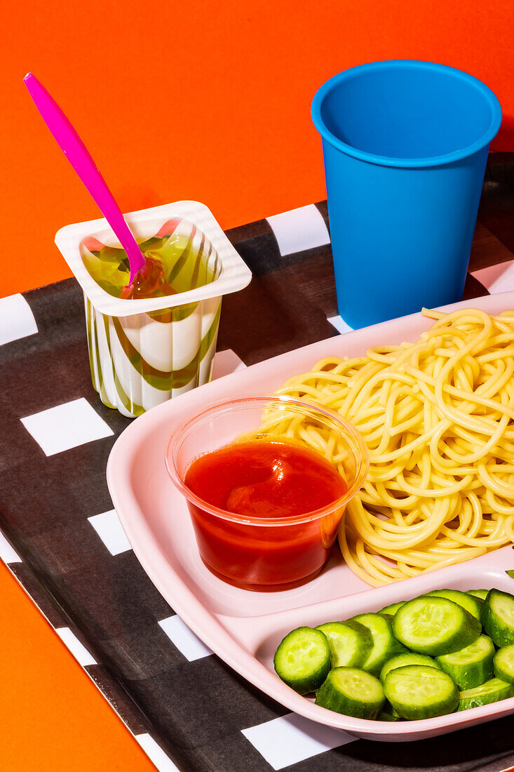 Spaghetti mit Tomatensoße von oben, frische Gurkenscheiben mit Löffel in einem Fruchtgummi-Becher und ein leeres Wasserglas, das zum Schulessen serviert wird