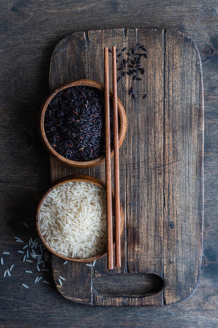 Draufsicht auf rohen schwarzen Wildreis und geschälten weißen Reis in den Schalen mit Stäbchen auf dem Schneidebrett auf dem Holztisch