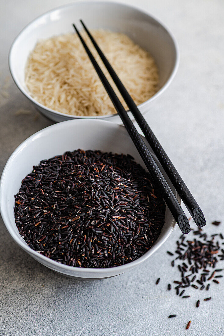 High angle of Raw wild black rice and peeled white rice in the bowls with chopsticks against white surface