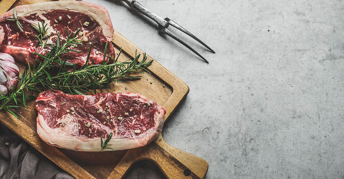 Raw beef steaks background with rosemary and green pepper on wooden cutting board with butcher fork. Close up. Preparing fresh meat. Top view with copy space.
