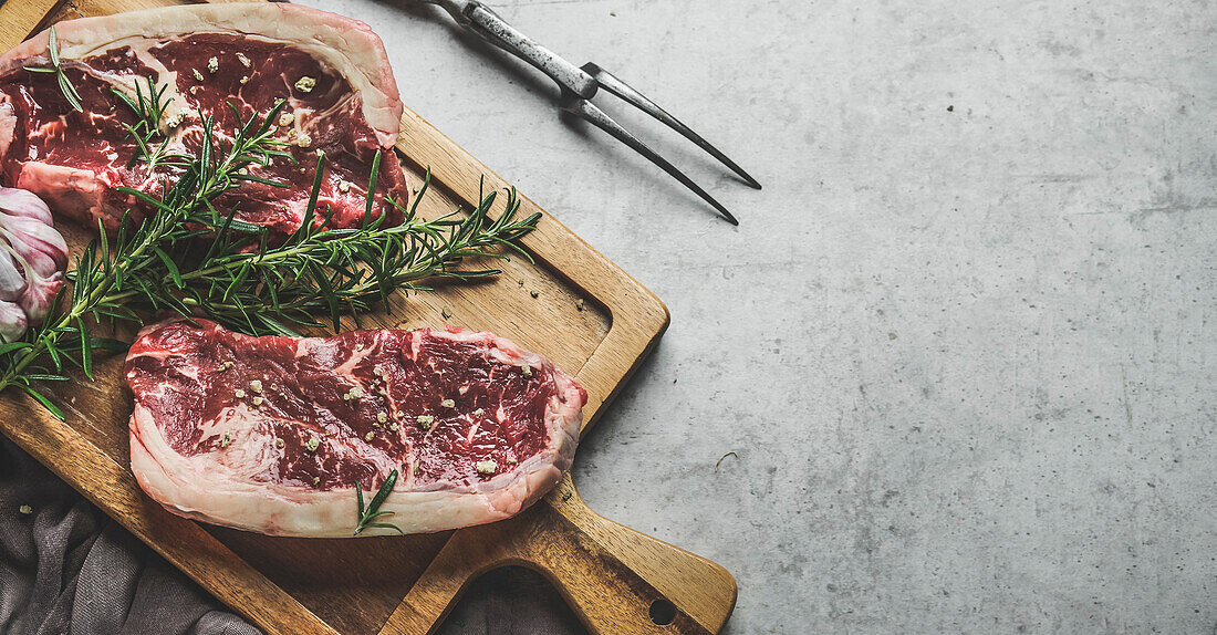 Rohe Rindersteaks mit Rosmarin und grünem Pfeffer auf einem hölzernen Schneidebrett mit einer Metzgergabel. Nahaufnahme. Vorbereitung von frischem Fleisch. Draufsicht mit Kopierbereich