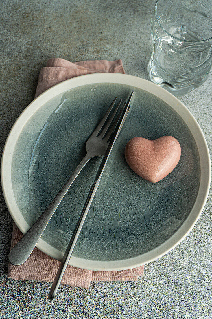 Draufsicht auf einen Keramikteller mit Besteck und Serviette mit herzförmigem Kristalldekor auf einer Betonfläche am Küchentisch für eine Mahlzeit während der Valentinstagsfeier