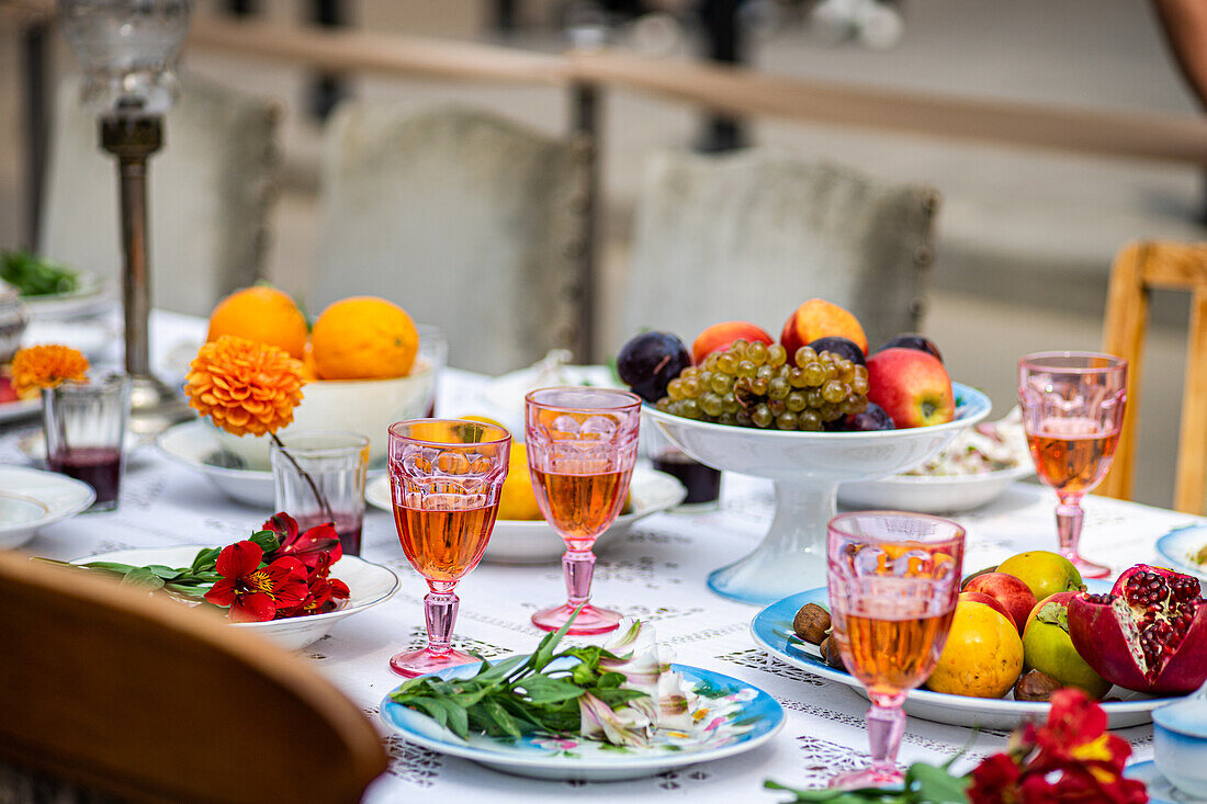 A vibrant outdoor table setting featuring an assortment of fresh fruits, colorful glasses filled with drinks, and decorative flowers placed on a white tablecloth.