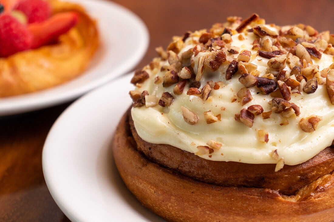Focus on an appetizing homemade cake with sweet cream and nuts on a white ceramic plate on a wooden dining table with other cakes in a blurred background indoors