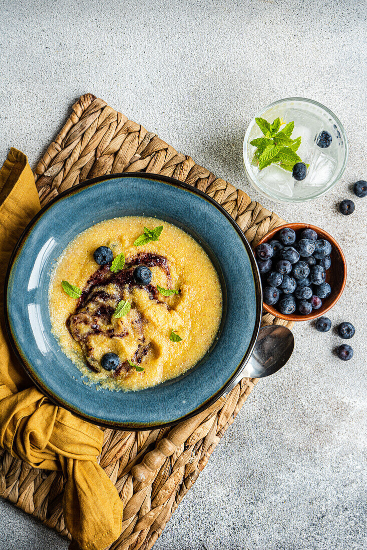 Top view of delicious traditional Italian polenta porridge topped with jam and mint leaves and served on table with bowl of fresh blueberries and glass of cold lemonade during breakfast